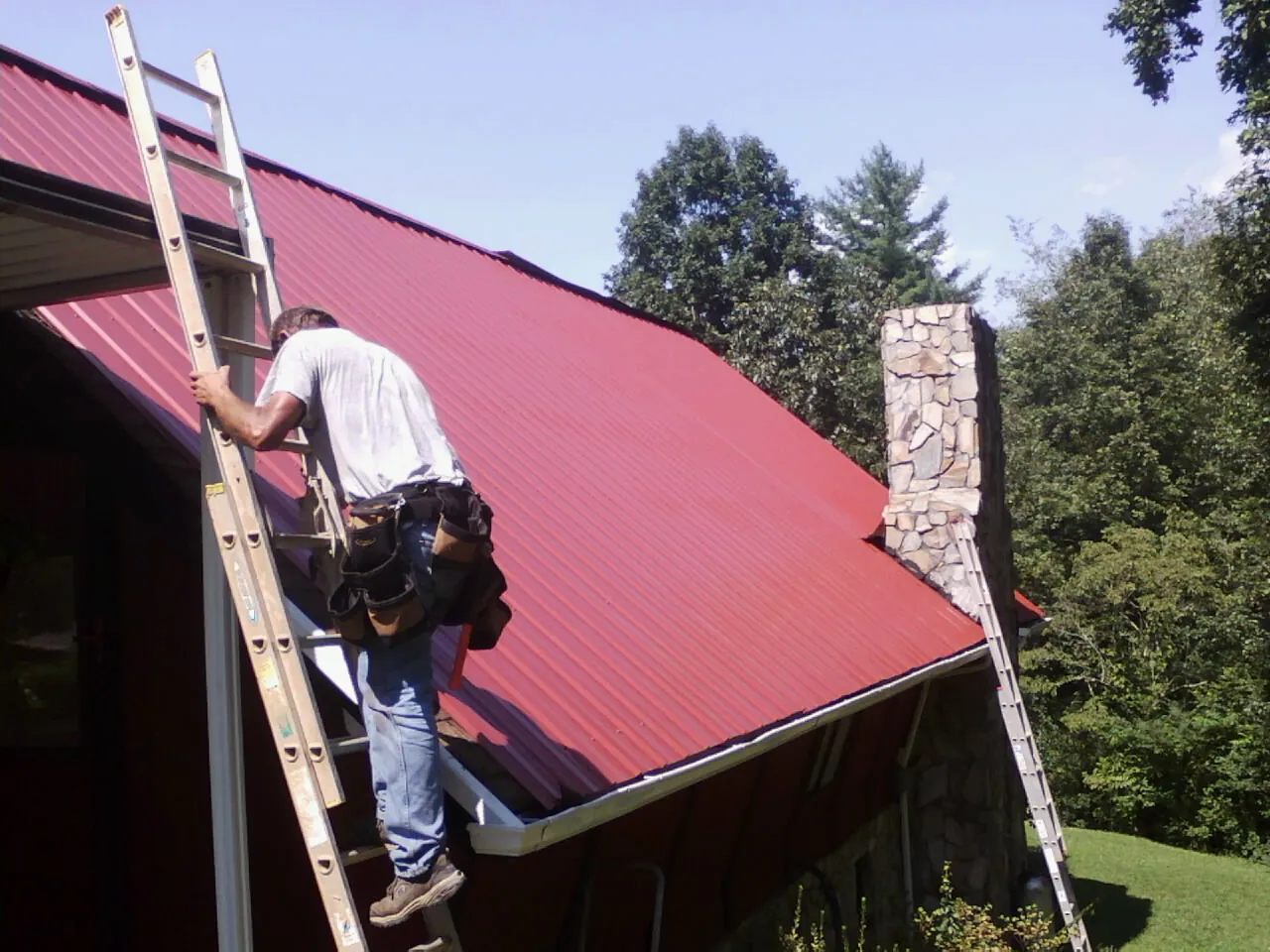 Roofer on a ladder beside a red metal roof and stone chimney — Soffit & Fascia Repair by Roofing Brothers in Leonardtown